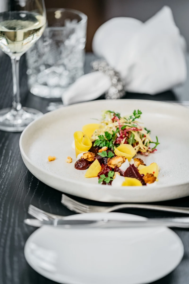 Elegantes Gericht auf weißem Teller mit gelben und roten Elementen, garniert mit frischen Kräutern. Ein Glas Weißwein und gestapelte weiße Teller im Hintergrund deuten auf ein gehobenes Restaurant oder Dining-Erlebis hin.