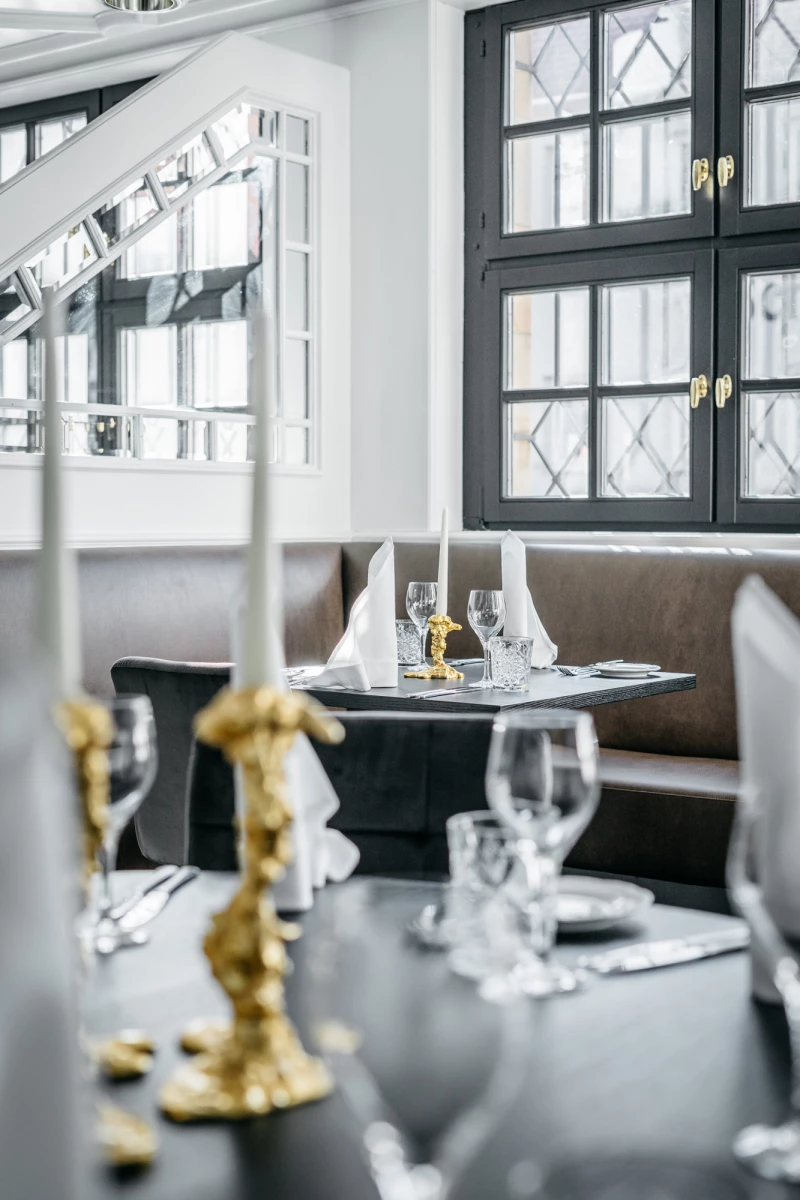 Gedeckter Tisch in elegantem Restaurant mit weißen Tellern, Weingläsern und goldenen Kerzenhaltern. Großes Fenster mit Sprossenfenstern im Hintergrund, gedämpftes Licht und stilvolle Einrichtung.