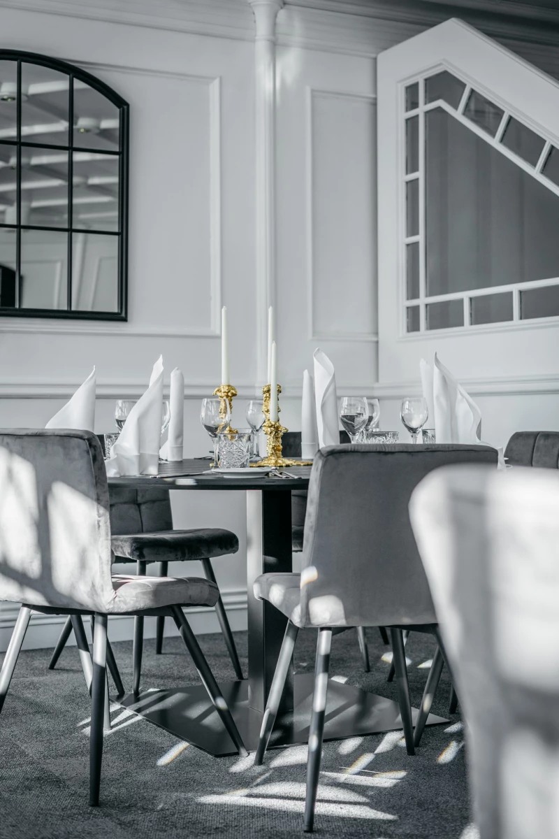 Eleganter Esszimmerbereich mit gedecktem Tisch, weißen Stoffservietten und goldenen Kerzenhaltern. Moderne graue Stühle stehen um einen Tisch, umgeben von stilvollen weißen Wänden und großen Fenstern.