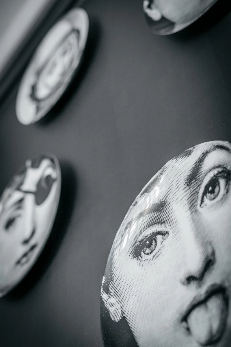 Mehrere schwarzweiße Teller oder Keramikscheiben an der Wand, die Gesichtsteile mit detaillierten Augen und Gesichtskonturen zeigen. Die Teller sind in unterschiedlicher Unschärfe und Perspektive angeordnet, was einen künstlerischen und surrealen Eindruck vermittelt.
