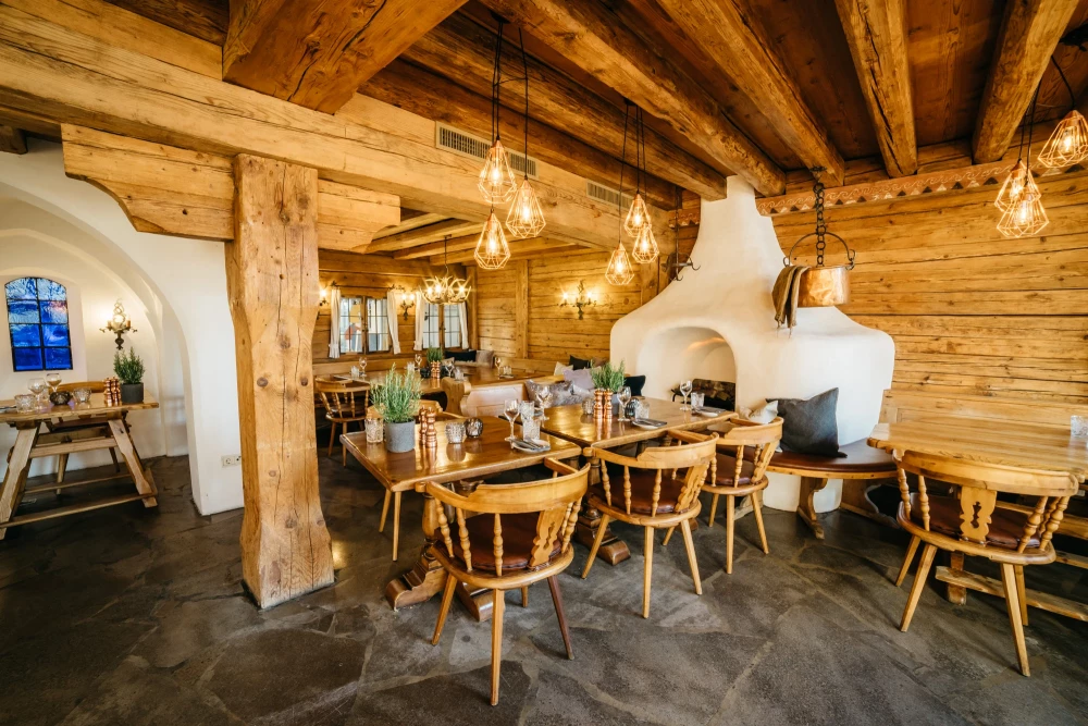 Gemütlicher Gastraum in Holzblockhaus-Optik mit rustikalen Holzbalken, hängenden Pendelleuchten und gedeckten Holztischen mit Stühlen. Ein weiß gefliester Kachelofen und Holzwände schaffen eine warme, einladende Atmosphäre in traditionellem Bergstil.