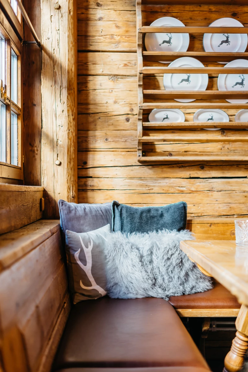 Gemütliche Holzstube mit rustikaler Sitzbank, weichen Kissen in Blau- und Grautönen und einem Tellerregal an der Holzwand. Warmes Sonnenlicht fällt durch ein Holzfenster und beleuchtet den einladenden, traditionellen Wohnbereich.