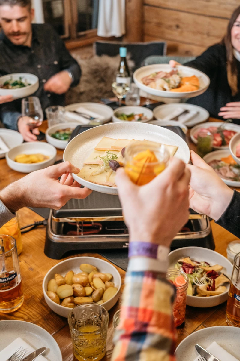Gemütliche Raclette-Mahlzeit mit verschiedenen Tellern, Beilagen und Zutaten auf einem Holztisch. Eine Hand hält eine Pfanne mit geschmolzenem Käse und einer Orangenscheibe, während im Hintergrund weitere Personen am Tisch sitzen.