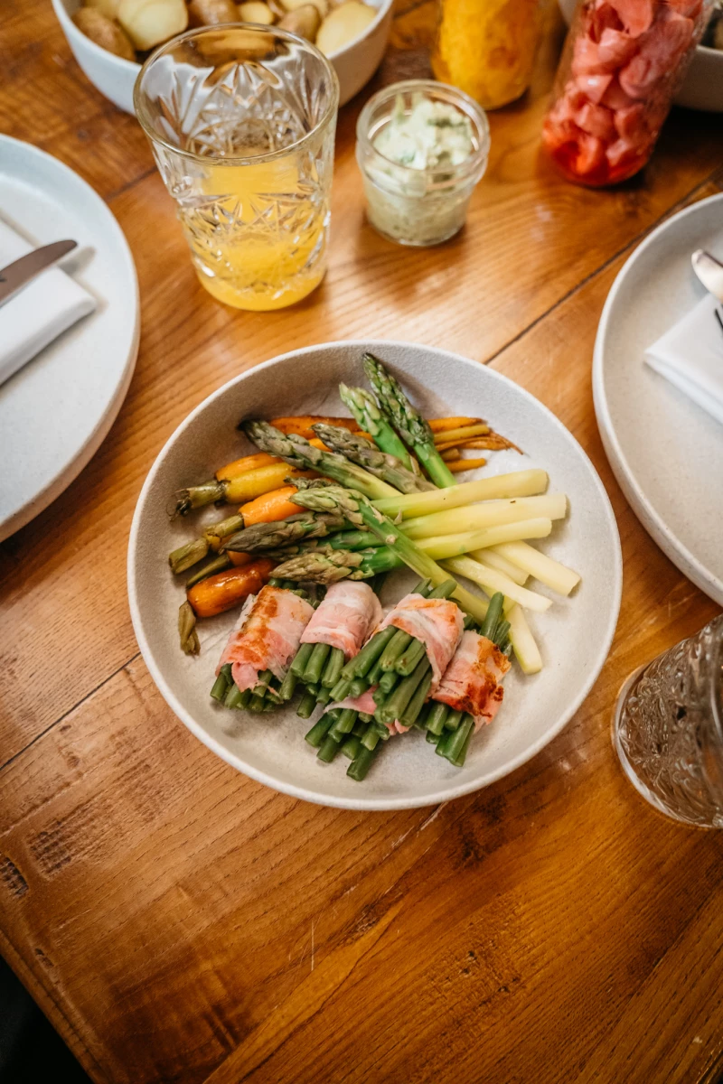 Gedämpfter Spargel, umwickelt von Schinken, serviert auf einem weißen Teller, daneben gelbes Getränk und Dekoration auf einem rustikalen Holztisch. Verschiedene Gemüsesorten wie grüner und gelber Spargel sowie Karotten sind appetitlich angerichtet.