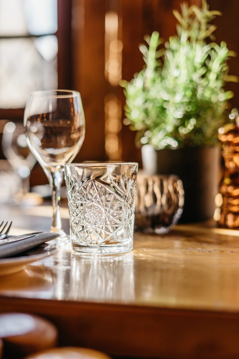 Geschliffenes Whiskyglas auf einem goldenen Holztisch mit unscharfem Hintergrund, im Hintergrund eine kleine grüne Pflanze in einem Topf.