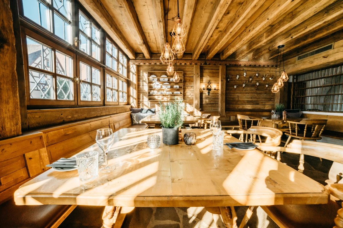 Rustikaler Gastraum mit Holzbalkendecke und großen Fenstern, an einem langen Holztisch gedeckt mit Gläsern und einer Zimmerpflanze. Vintage Hängelampen und warmes Sonnenlicht schaffen eine gemütliche Atmosphäre im Landhausstil.
