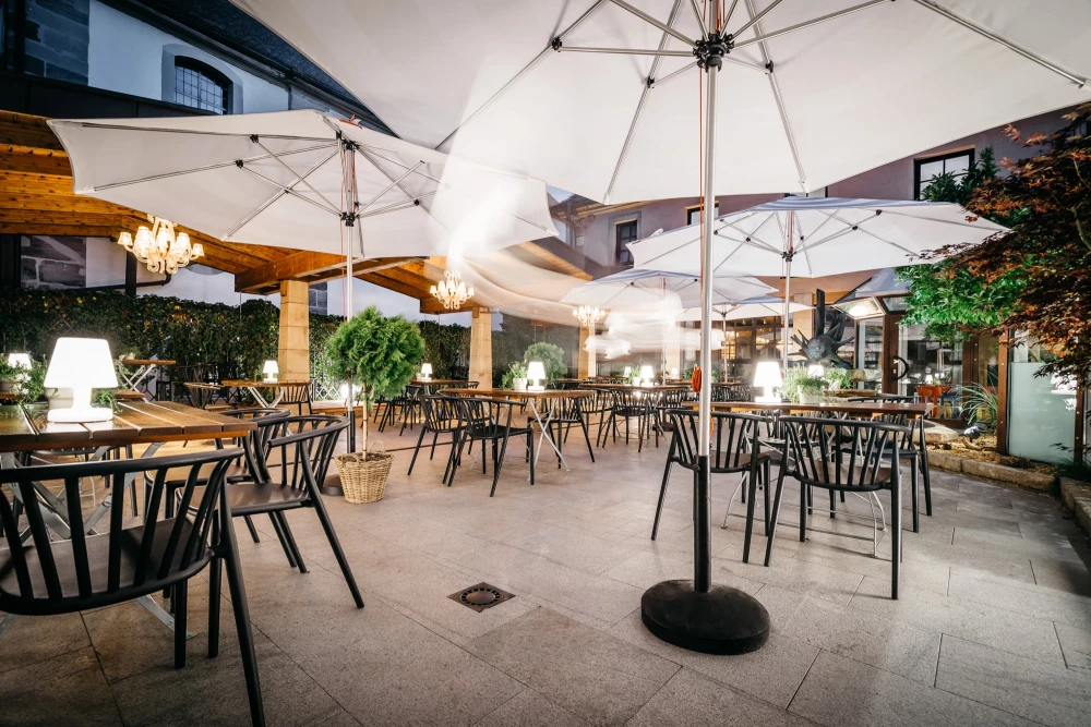 Heller Biergarten mit weißen Sonnenschirmen und schwarzen Metallstühlen, umgeben von Grünpflanzen und dekorativen Leuchten. Der gepflasterte Außenbereich bietet eine einladende Atmosphäre für Gäste zum Entspannen und Genießen.