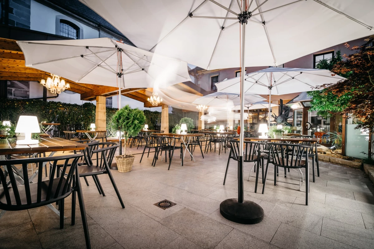Heller Biergarten mit weißen Sonnenschirmen und schwarzen Metallstühlen, umgeben von Grünpflanzen und dekorativen Leuchten. Der gepflasterte Außenbereich bietet eine einladende Atmosphäre für Gäste zum Entspannen und Genießen.