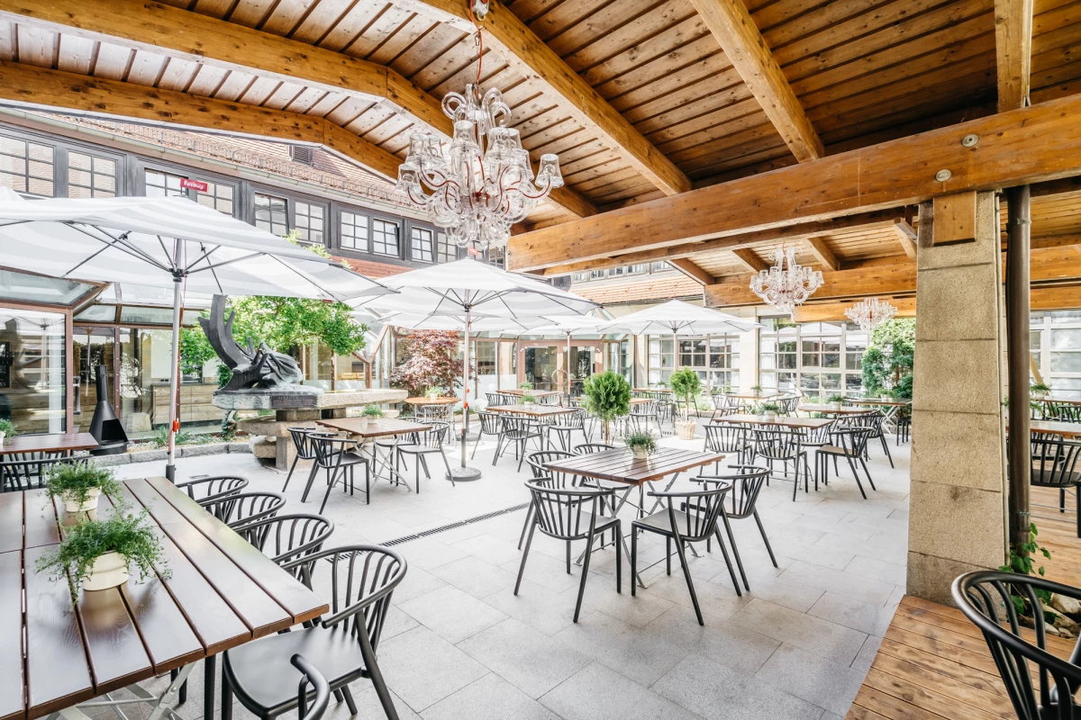 Geräumige Terrasse mit Holzdachstuhl, ausgestattet mit schwarzen Metalltischen und -stühlen sowie weißen Sonnenschirmen. Kristalllüster hängt von der Holzdecke, und die Einrichtung wirkt hell und einladend mit Blick auf einen Gartenbereich.