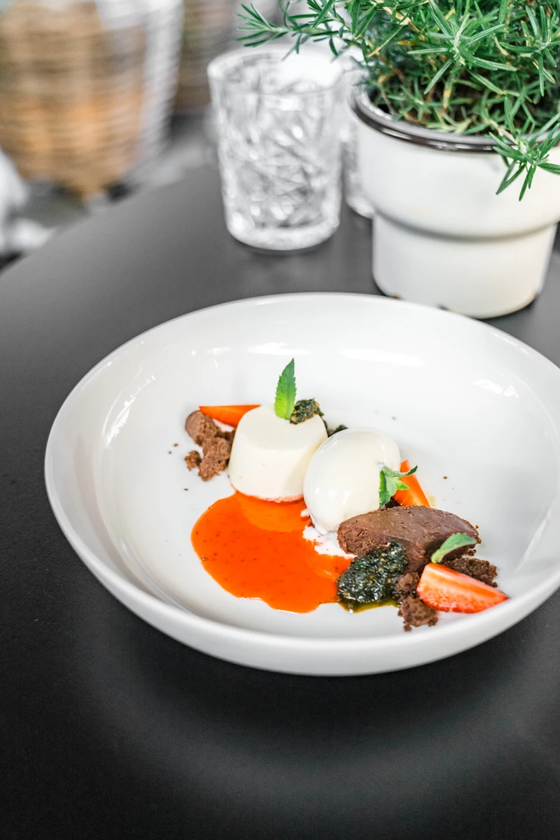 Raffiniertes Gourmet-Gericht auf weißem Teller mit zart angerichtetem Fleisch, cremigem weißem Klecks, orangefarbener Soße und grünen Kräutern, im Hintergrund ein Rosmarin-Topf und ein strukturiertes Glas.