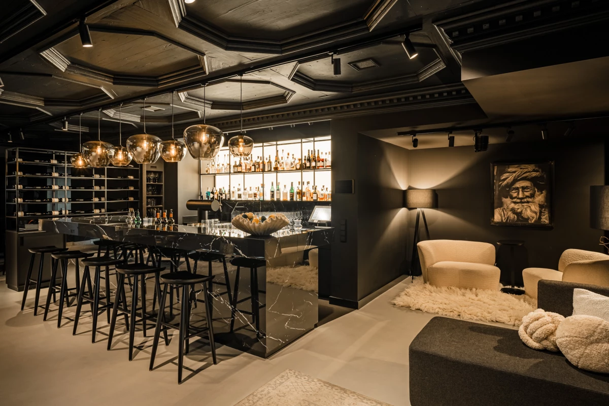 Stilvolle Bar mit elegantem schwarzem Tresen, goldenen Hängelampen und Barhocker. Rechts ein gemütlicher Sitzbereich mit beigen Polstermöbeln und einem Kunstportät an der Wand. Die Atmosphäre ist edel und modern mit gedämpftem Licht und hochwertigem Interieur.