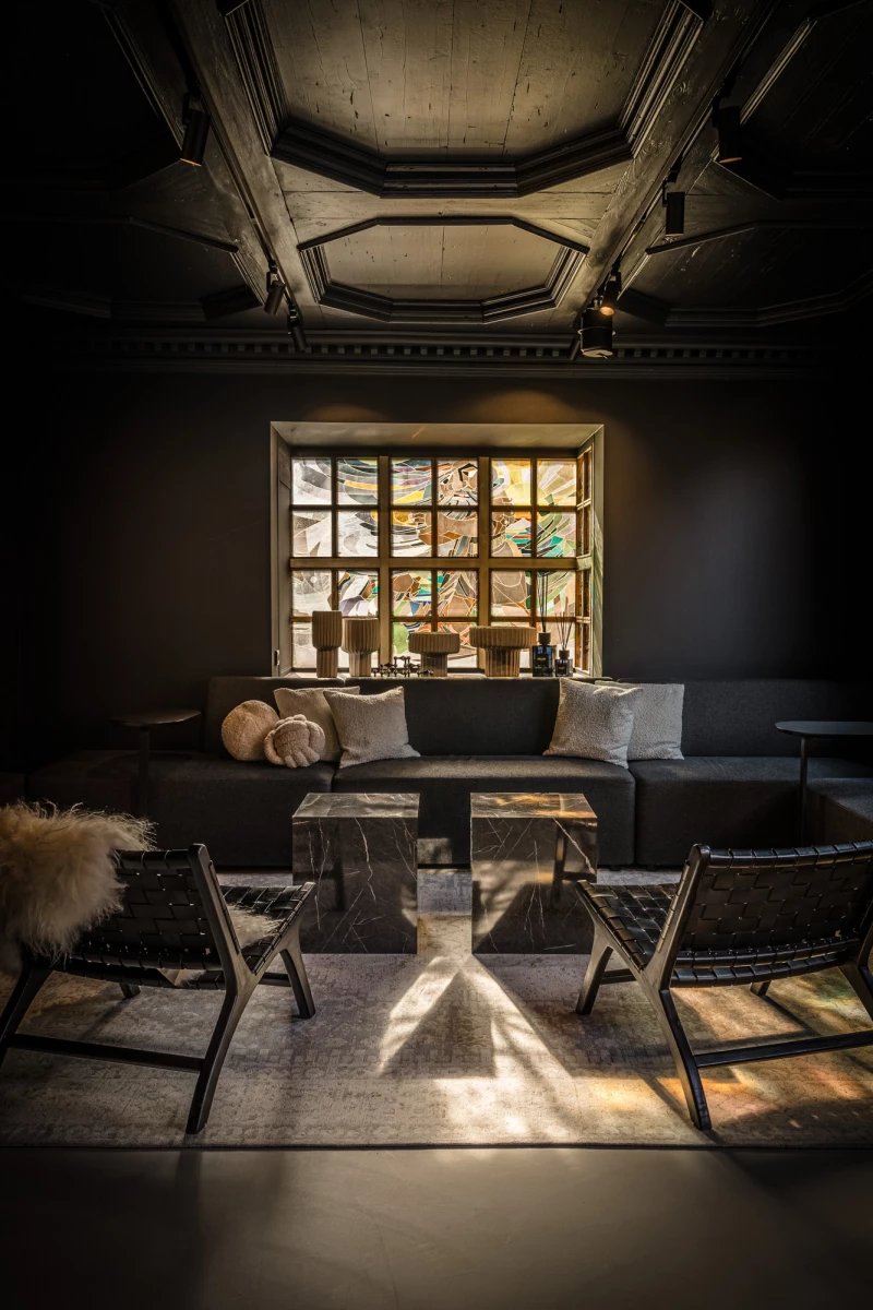 Rustikale Lounge mit Holzmöbeln und großem Fenster, das Tageslicht hereinlässt. Dunkle Lederstühle und Holztisch bilden einen gemütlichen Sitzbereich vor dem Fenster mit Ausblick auf Bäume.