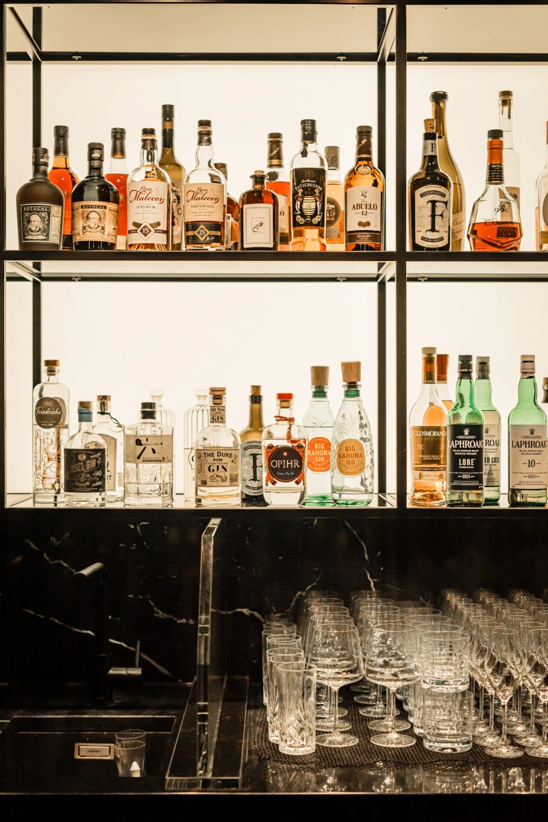 Elegante Hausbar mit zwei Regalbrettern voller verschiedener Spirituosen und Flaschen in warmen Tönen. Darunter eine Theke mit gestapelten Whiskygläsern und Kristallgläsern. Die Anordnung ist symmetrisch und ordentlich, mit einer Vielzahl von Alkoholen in unterschiedlichen Formen und Farben.