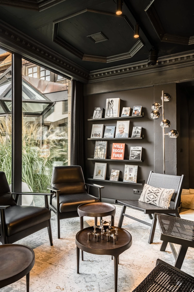 Stilvoller Lesebereich mit Panoramafenstern und schwarzen Wänden, ausgestattet mit Ledersesseln, Couchtischen und einer Kunstwand mit sorgsam arrangierten Büchern und Bildern. Dezente Beleuchtung und ein modernes, minimalistisches Design schaffen eine einladende Atmosphäre.