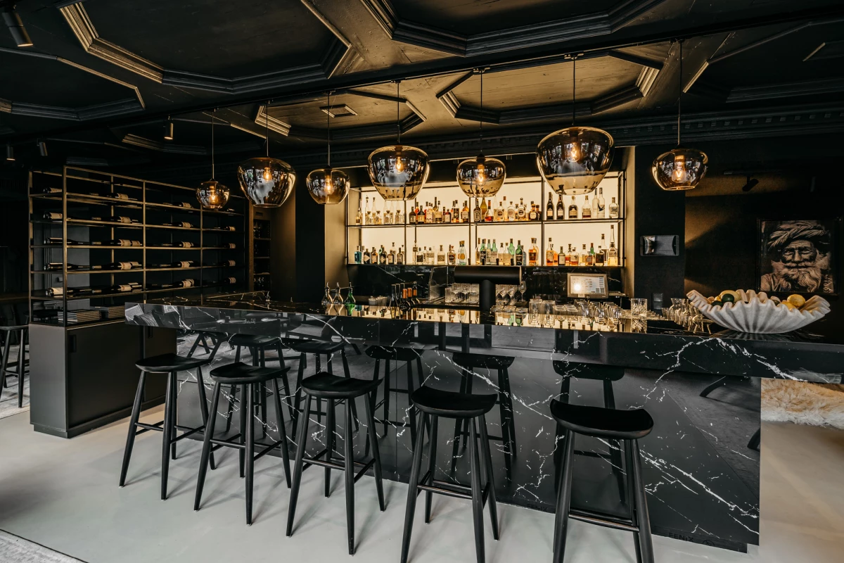 Stilvolle Bar mit elegantem, schwarzem Marmor-Tresen und hohen Barhockern, goldenen Hängelampen und einer beeindruckenden Spirituosensammlung an der Rückwand. Die Decke ist aufwendig gestaltet und der Raum strahlt eine moderne, luxuriöse Atmosphäre aus.