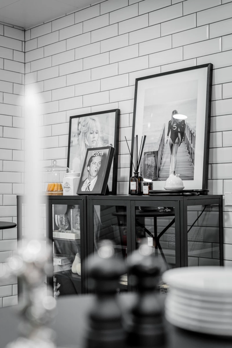 Schwarzweißes Stillleben einer Küchen-Sideboard-Ecke mit gerahmten Kunstdrucken und Dekorationsobjekten vor einer weißen Ziegelmauer. Auf dem Sideboard stehen verschiedene Bilderrahmen, Kerzen und Dekoration, im Vordergrund sind unscharf Geschirr und Gegenstände zu sehen.
