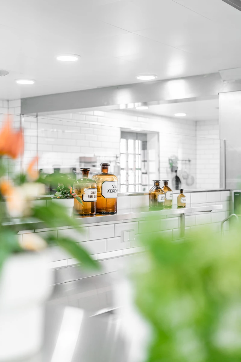 Bernsteinfarbene Apothekergläser stehen in einer weißen Küche mit weißen Wandfliesen und Deckenbeleuchtung. Im Vordergrund verschwimmen grüne Blätter und Blumen, während die Glasflaschen scharf im Fokus sind. Die helle, moderne Kücheneinrichtung strahlt Sauberkeit und Eleganz aus.