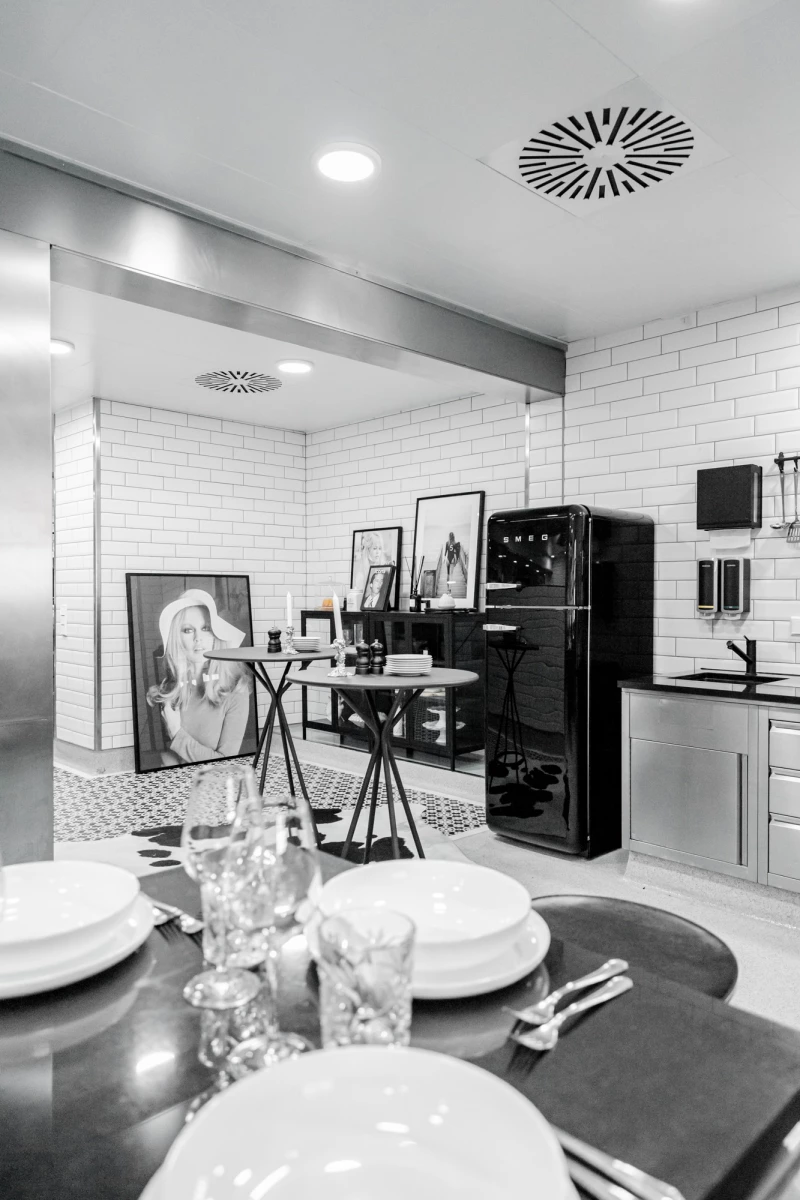 Moderne Küche mit schwarzer Retro-Kühlschrank, Ziegelfliesen und gedecktem Tisch im Vordergrund. Kunstvolle Atmosphäre mit Gemälde und Rahmen, die an einer Wand lehnen. Dezentes Licht und klare Linien prägen den minimalistischen Raum.