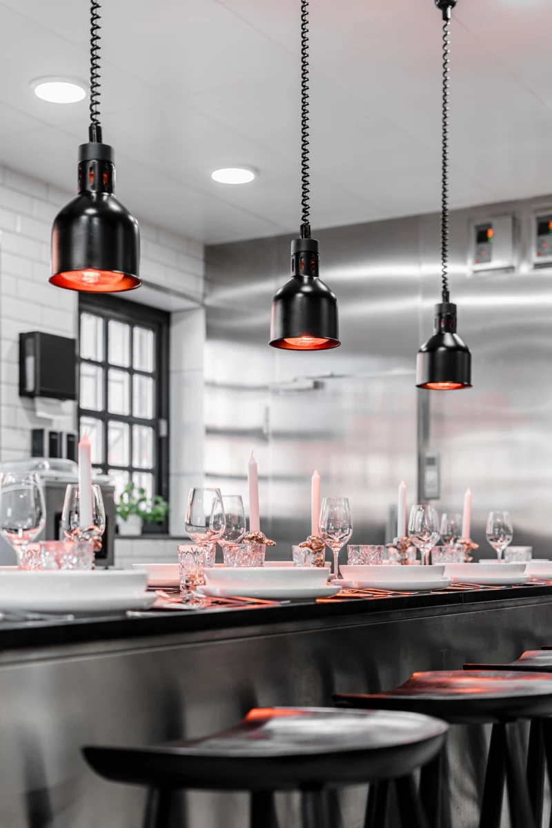 Moderne Bar-Theke mit schwarzen Hängelampen und rosa Weingläsern, dezent beleuchtet und elegant eingerichtet. Stahlgraue Oberflächen und große Fenster verleihen dem Raum eine zeitgenössische Atmosphäre.