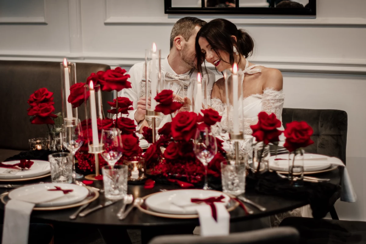 Festlich gedeckter Tisch mit roten Rosen und brennenden Kerzen, zwei Personen in Hochzeitskleidung sitzen nah beieinander und teilen einen intmen Moment an einem eleganten Esstisch mit weißem Geschirr und Kristallgläsern.