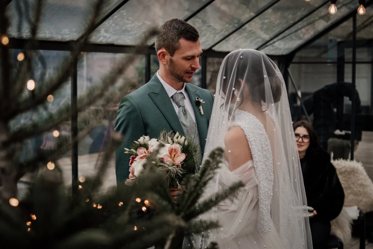 Brautpaar in einem Wintergarten mit Lichterketten, der Bräutigam in einem grünen Anzug und die Braut in einem weißen Kleid mit Schleier, während sie einen Blumenstrauss hält.