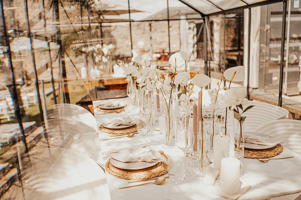 Gedeckter Tisch in einem Wintergarten oder Gewächshaus mit weißer Tischdecke, goldenen Platztellern und eleganten Glasvasen mit weißen Blumen. Die Tischdekoration ist minimalistisch und zeitlos, mit einer warmen, einladenden Atmosphäre.