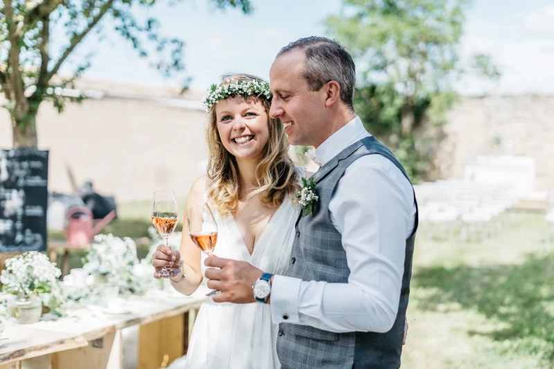 Braut mit Blumenkranz und Bräutigam in Weste stehen an einem Hochzeitstag auf einer Wiese, halten Sektgläser und lächeln glücklich miteinander.