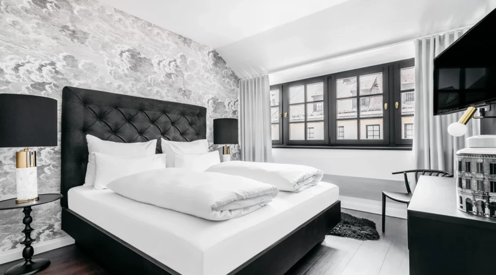 Ein elegant eingerichtetes Hotelzimmer mit einem schwarzen gepolsterten Bett, weißer Bettwäsche und einem großen Fenster. Graue Tapete und schwarze Akzente wie Nachttischlampen und Bodenbelag verleihen dem Raum einen modernen, kontrastierenden Look.