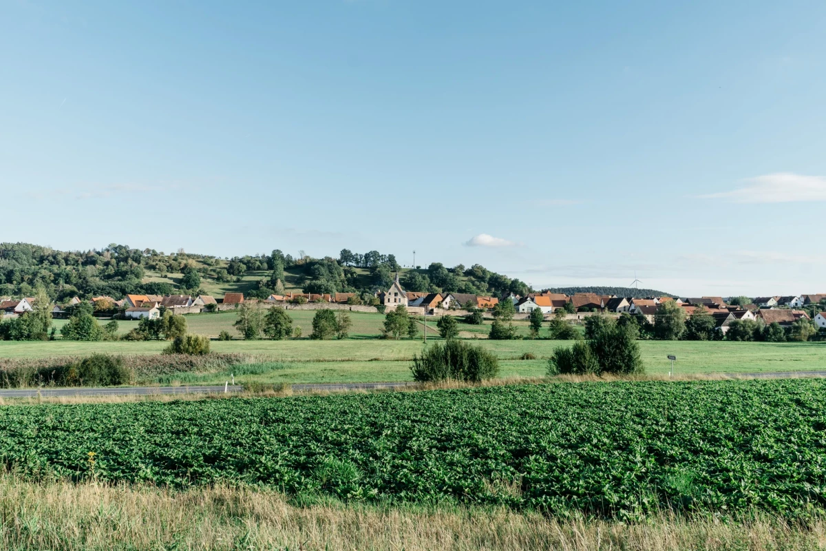 Grüne Landschaft mit Feldern im Vordergrund und einem Dorf mit roten Dächern am Horizont. Sanfte Hügel und blauer Himmel bilden eine friedliche ländliche Szene.