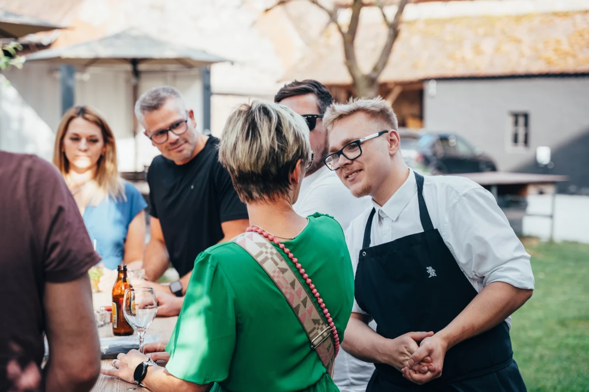 Gruppe von Menschen unterhält sich auf einem Hoffest, ein junger Mann in Schürze steht im Vordergrund und lächelt, während andere Personen im Hintergrund Bier trinken und plaudern.