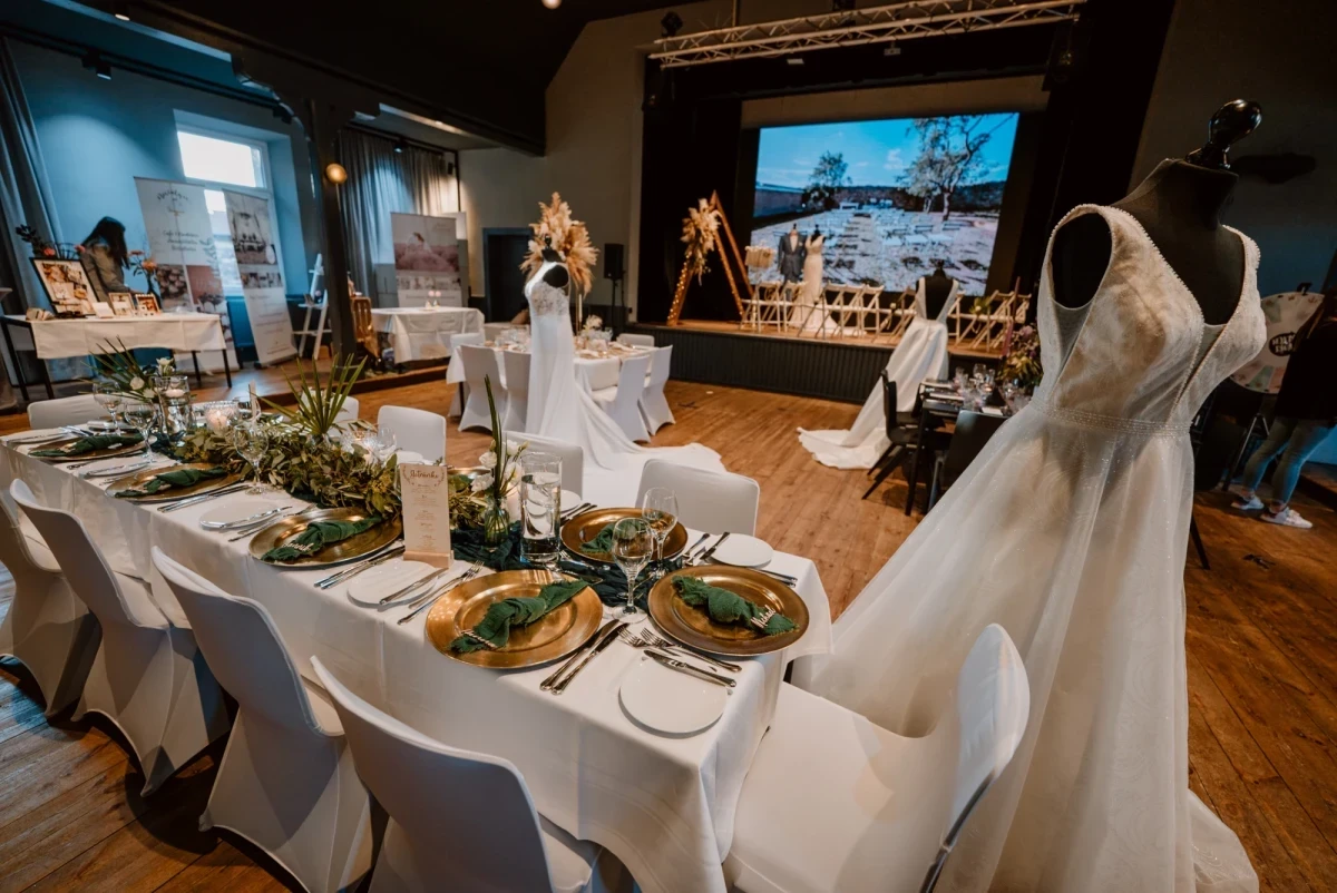 Hochzeitstafel mit weiß gedeckten Tischen, goldenen Tellern und Gründekor, einem Brautkleid auf einer Schaufensterpuppe im Vordergrund und einer Präsentation auf einer Leinwand im Hintergrund, die eine Hochzeitsveranstaltung in einem Veranstaltungsraum zeigt.