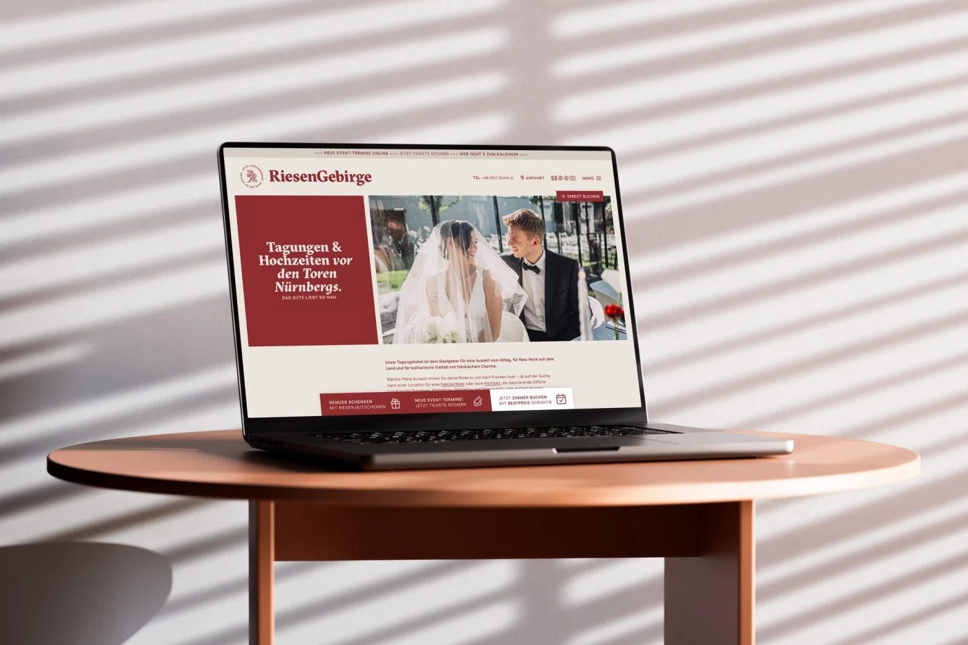 Laptop auf einem runden Holztisch, der die Website von Hotel Riesengebirge präsentiert, vor einer Wand mit Lichtschatten-Streifen.