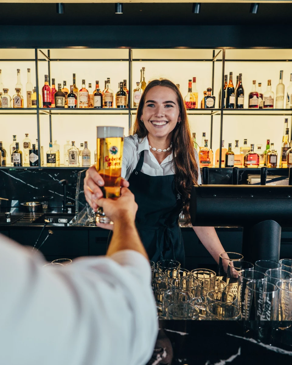 Eine lächelnde Barkeeperin in Schürze reicht ein Bierglas über die Theke, im Hintergrund eine Reihe von Spirituosenflaschen in einem Bar-Regal.