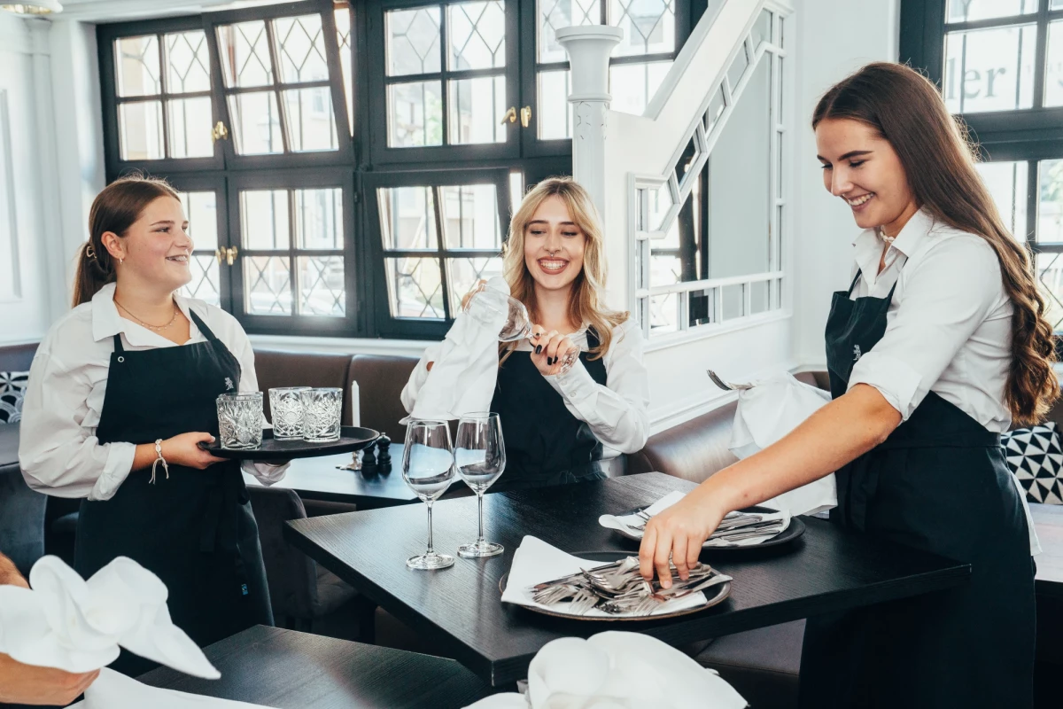 Drei Kellnerinnen in schwarzen Schürzen bereiten einen Tisch in einem hellen, modernen Restaurant vor. Sie lächeln und arbeiten gemeinsam mit Tellern und Weingläsern. Die große Fensterfront im Hintergrund lässt viel natürliches Licht in den Raum.