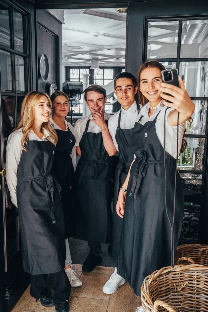 Fünf junge Mitarbeiter in grauen Schürzen stehen in einem modernen Café oder Restaurant und eine Person macht ein Selfie. Sie lächeln und posieren gemeinsam für ein Gruppenfoto, das die Teamstimmung und Arbeitsatmosphäre einfängt.