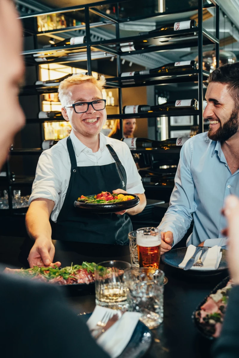 Ein Kellner mit Brille und schwarzer Schürze serviert ein buntes Gericht in einem Restaurant, während ein Gast lächelnd daneben steht und ein Bier hält.