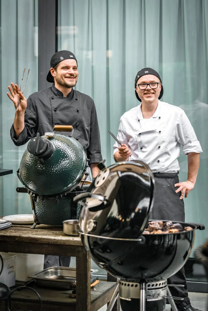 Zwei Köche in Berufskleidung stehen neben großen Grillgeräten, einer in grauer und einer in weißer Jacke, beide lächelnd und in einer entspannten Arbeitshaltung.