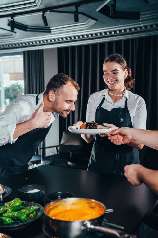 Ein Küchenchef in schwarzer Schürze und eine lächelnde Servicekraft in weißem Hemd und schwarzer Schürze bereiten gemeinsam ein Gericht in einer professionellen Restaurantküche zu.