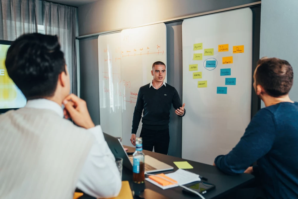 Ein Vortragender steht in einem Meetingraum vor einem Whiteboard. Zwei Personen sind von hinten zu sehen und blicken auf den Vortragenden.