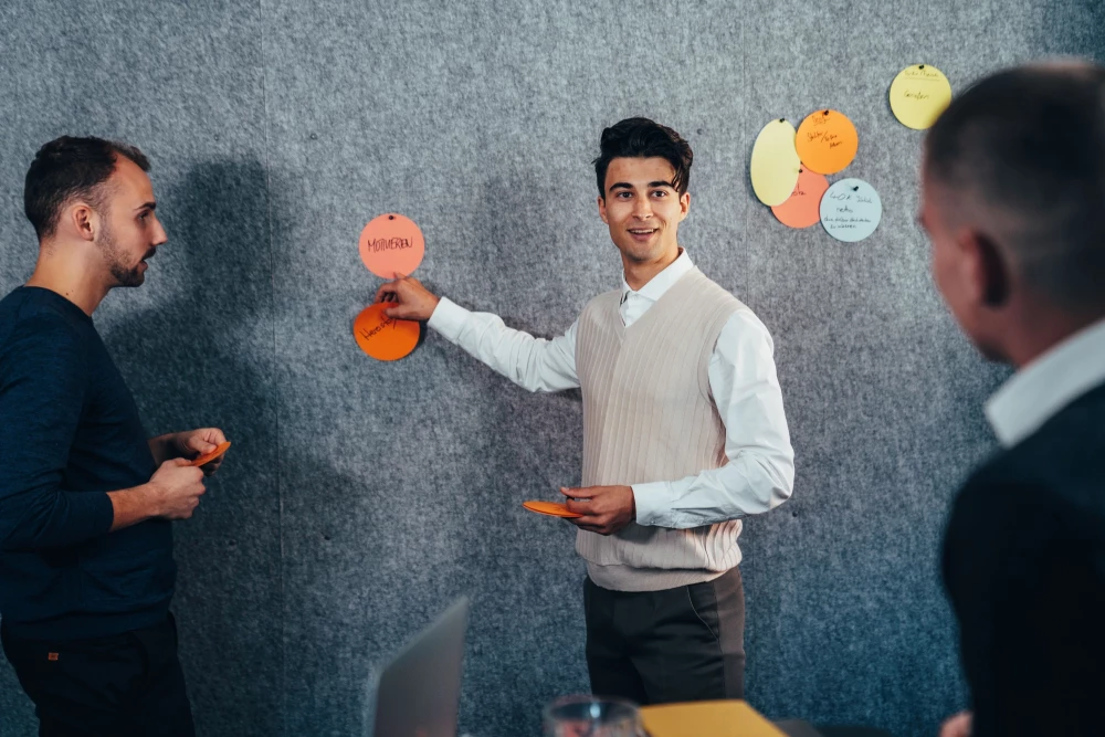 An einer Filzwand heftet ein Workshop-Teilnehmer Metaplankarten an und richtet dabei seine Aufmerksamkeit auf einen anderen Teilnehmer, der von hinten zu sehen ist. Ein weiterer Teilnehmer steht neben dem Teilnehmer im Fokus.