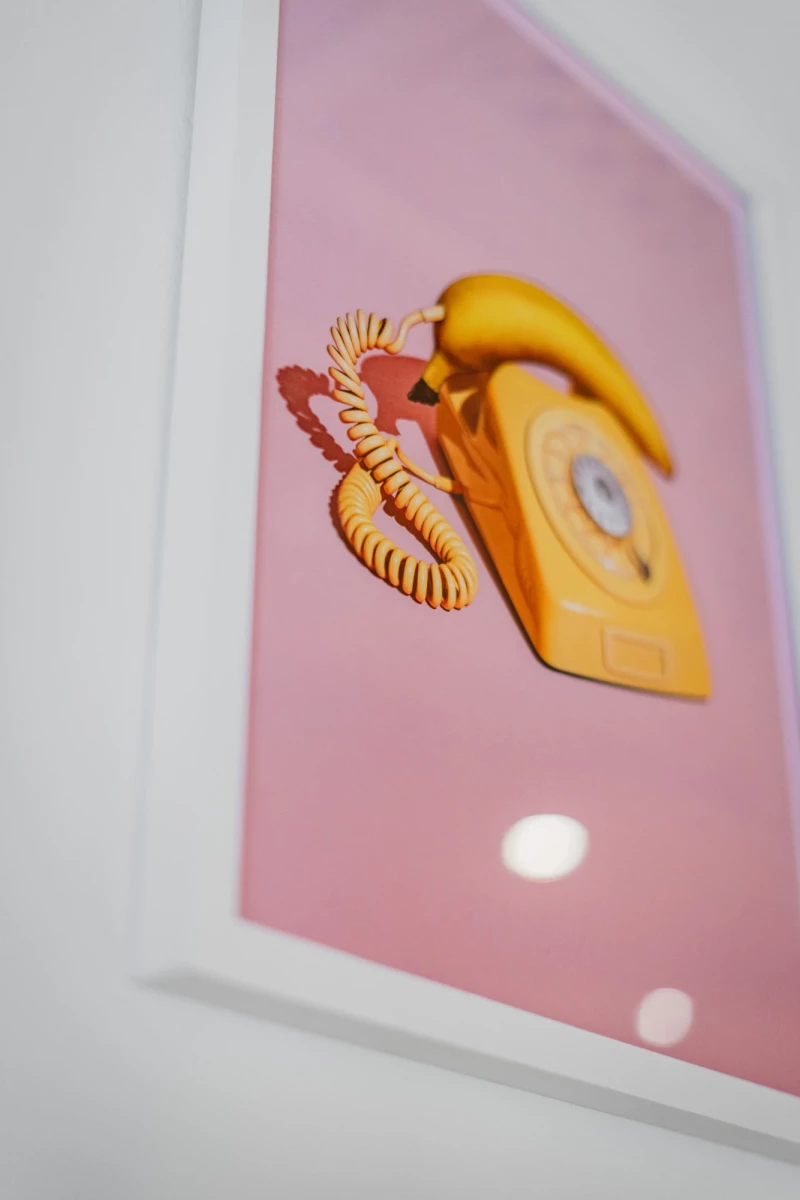 Gelbes Vintage-Telefon mit gedrehter Schnur auf einem rosa Hintergrund, das leicht schräg in einem weißen Rahmen positioniert ist und eine nostalgische, minimalistisch gestaltete Atmosphäre ausstrahlt.