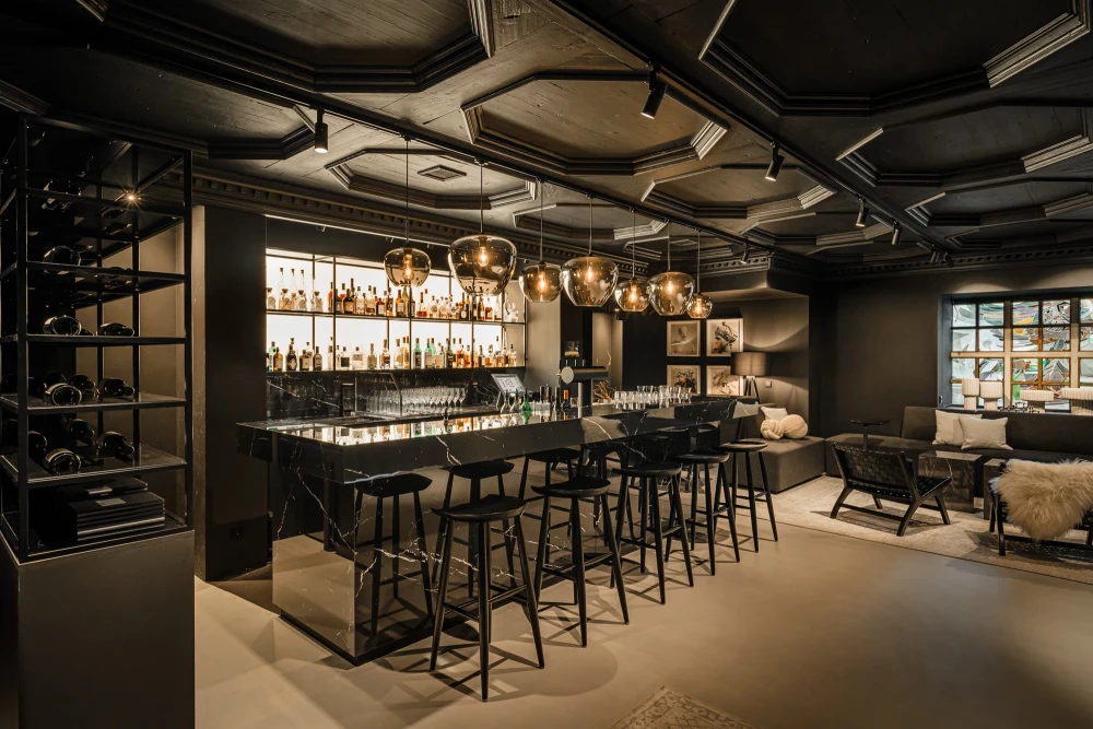 Moderner Bar-Bereich mit schwarzen Barhockern, goldenen Pendelleuchten und einer Regalwand voller Spirituosen. Die Decke ist geometrisch gestaltet, und im Hintergrund ist eine gemütliche Sitzecke mit einem Sessel zu sehen.