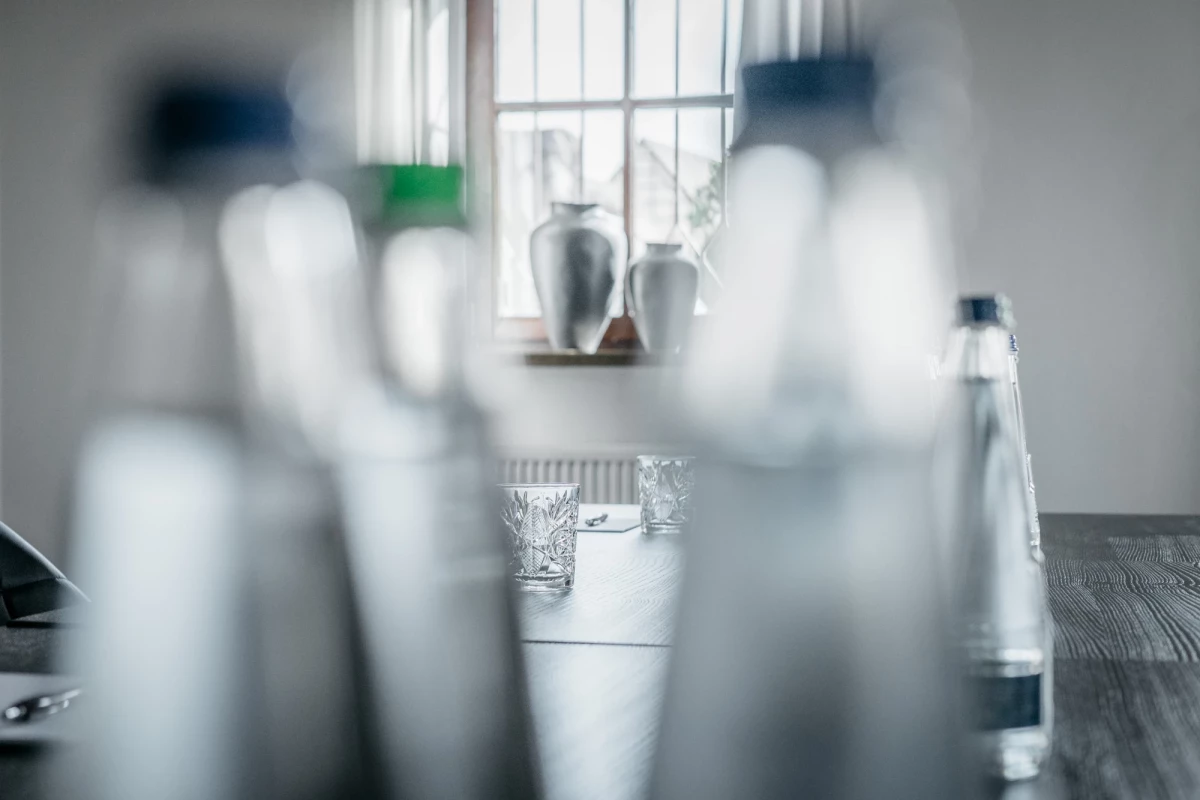 Mehrere unscharfe Glasflaschen im Vordergrund, im Hintergrund ein Fenster mit Vorhängen und einem Blumenstrauss. Ein Kristallglas steht auf einem Holztisch, der Raum wirkt gedämpft und minimalistisch.