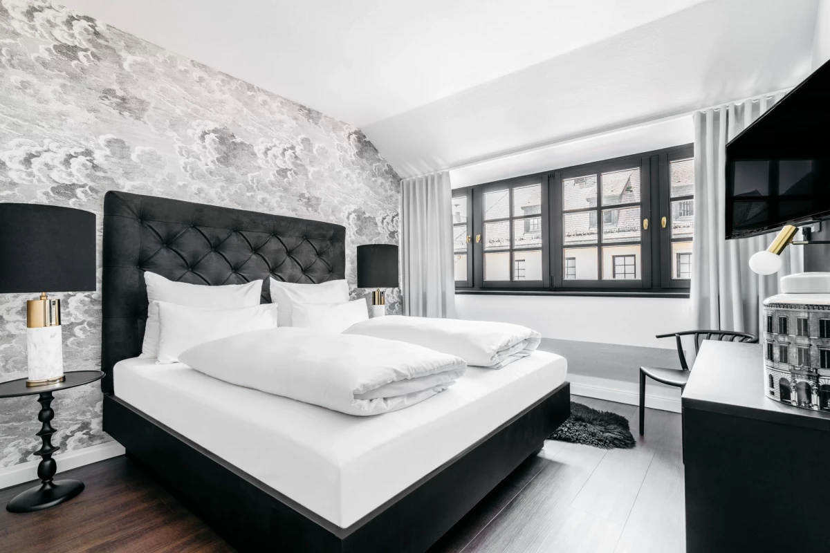 Modernes Hotelzimmer in Schwarz-Weiß mit gepolstertem Bett, gemusteter Akzentwand und großem Fenster. Dunkler Holzboden, weiße Bettwäsche und elegante schwarze Nachttischlampen komplettieren das stilvolle Interieur.