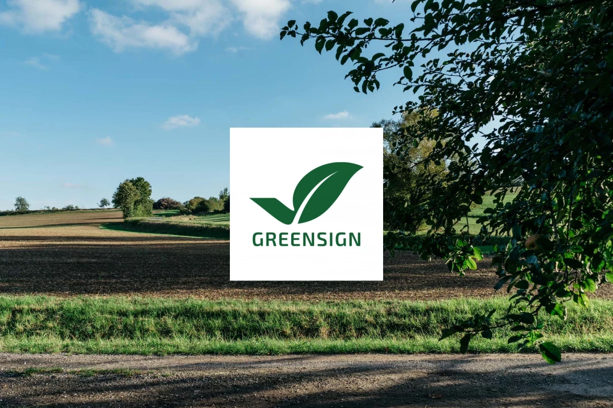 Grüne Landschaft mit Feldern, Bäumen und einem weißen Quadrat mit dem Greensign-Logo in der Mitte. Das Logo zeigt ein stilisiertes grünes Blatt mit einem Häkchen.
