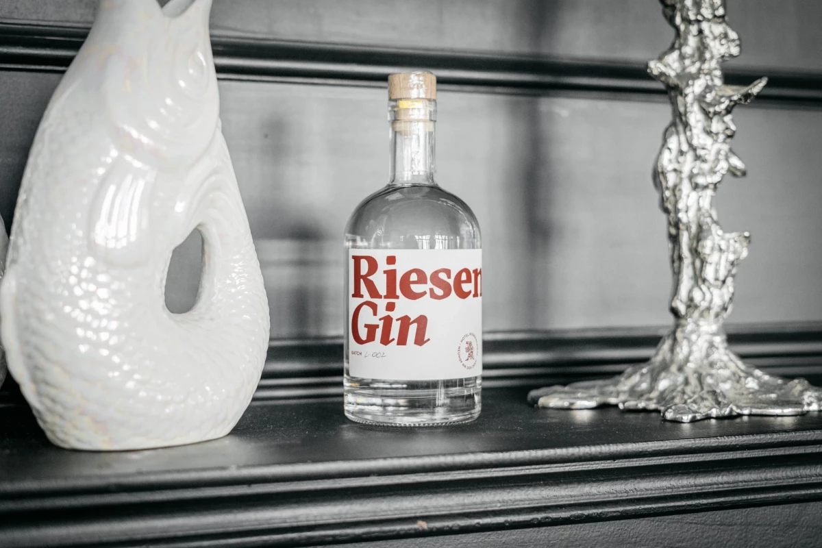 Flasche Rieser Gin auf einem dunklen Regal zwischen einer weißen Keramikvase und einem silbernen Kerzenhalter. Die Flasche hat ein klares Design mit rotem Schriftzug und Korken.