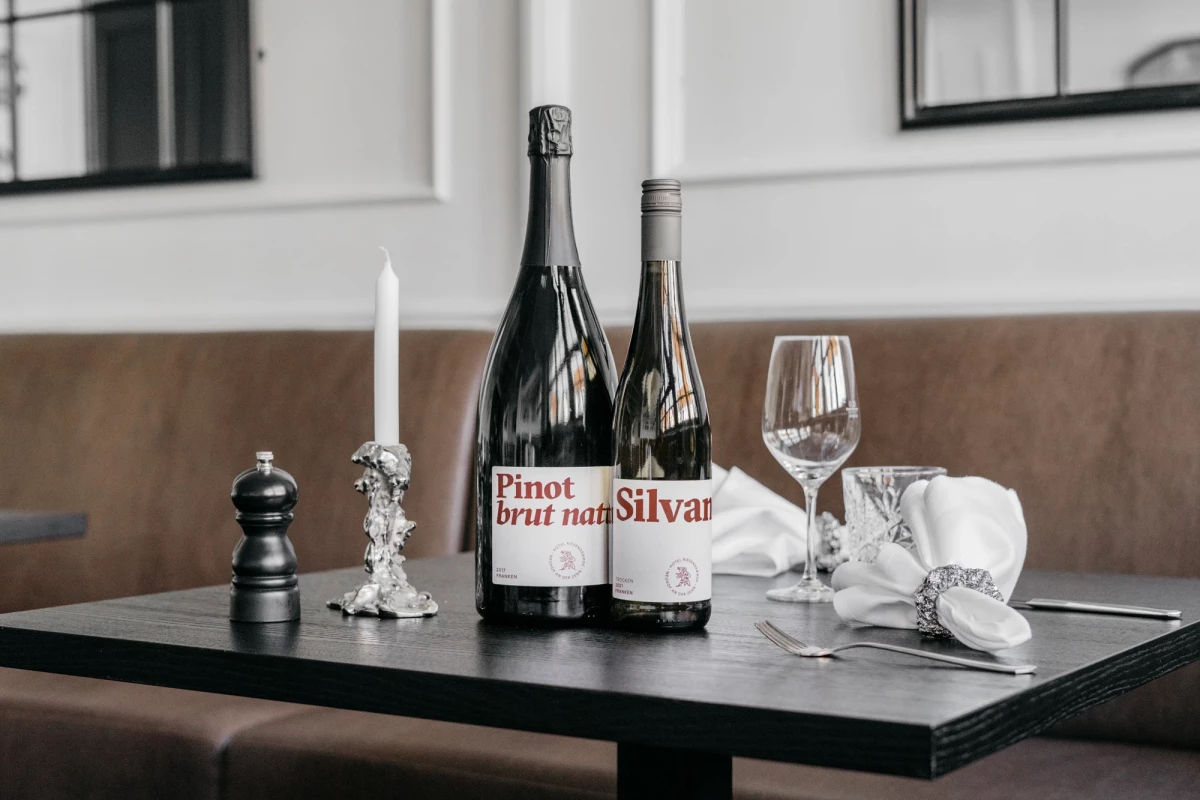 Zwei Weinflaschen von Pinot Brut nah und Silvar stehen auf einem dunklen Holztisch neben einem Weinglas, einer Kerze und einer Pfeffermühle in gedämpften Farbtönen.