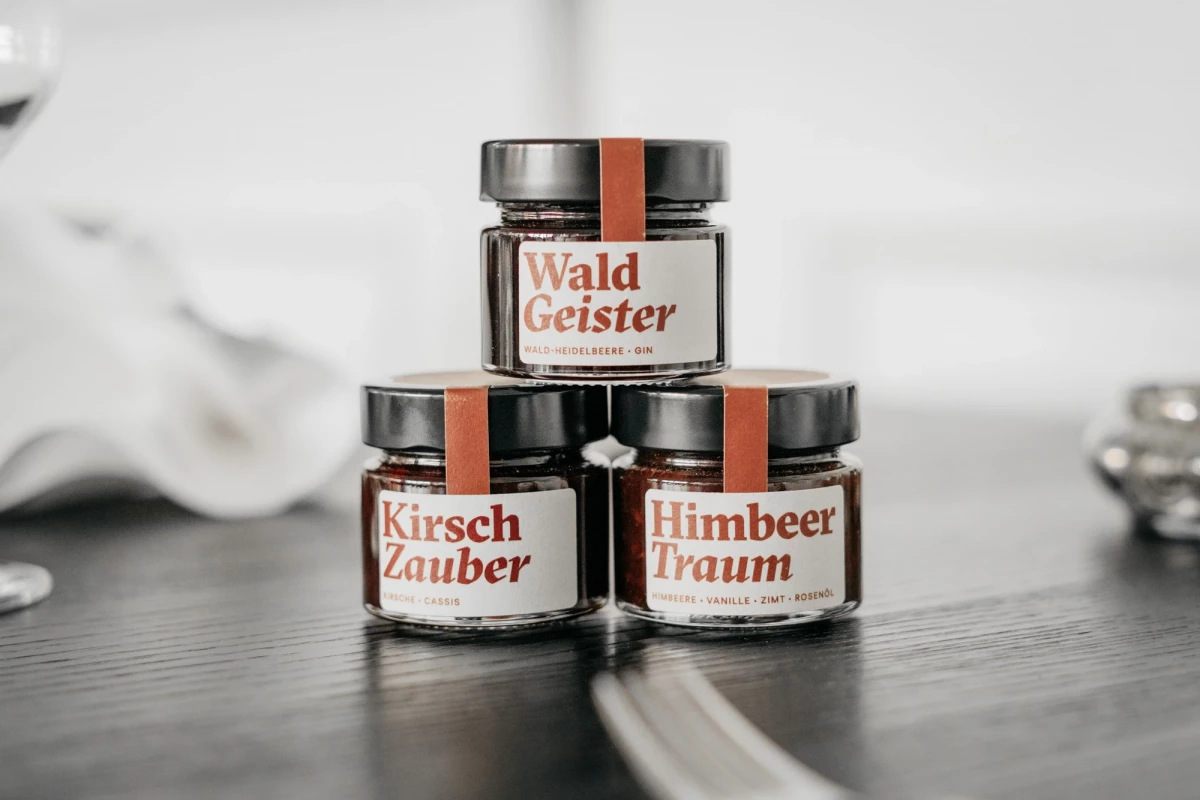 Drei Glasgefäße mit verschiedenen Etiketten von Wald Geister, Kirsch Zauber und Himbeer Traum, gestapelt auf einer dunklen Oberfläche in einem minimalistischen Setting.