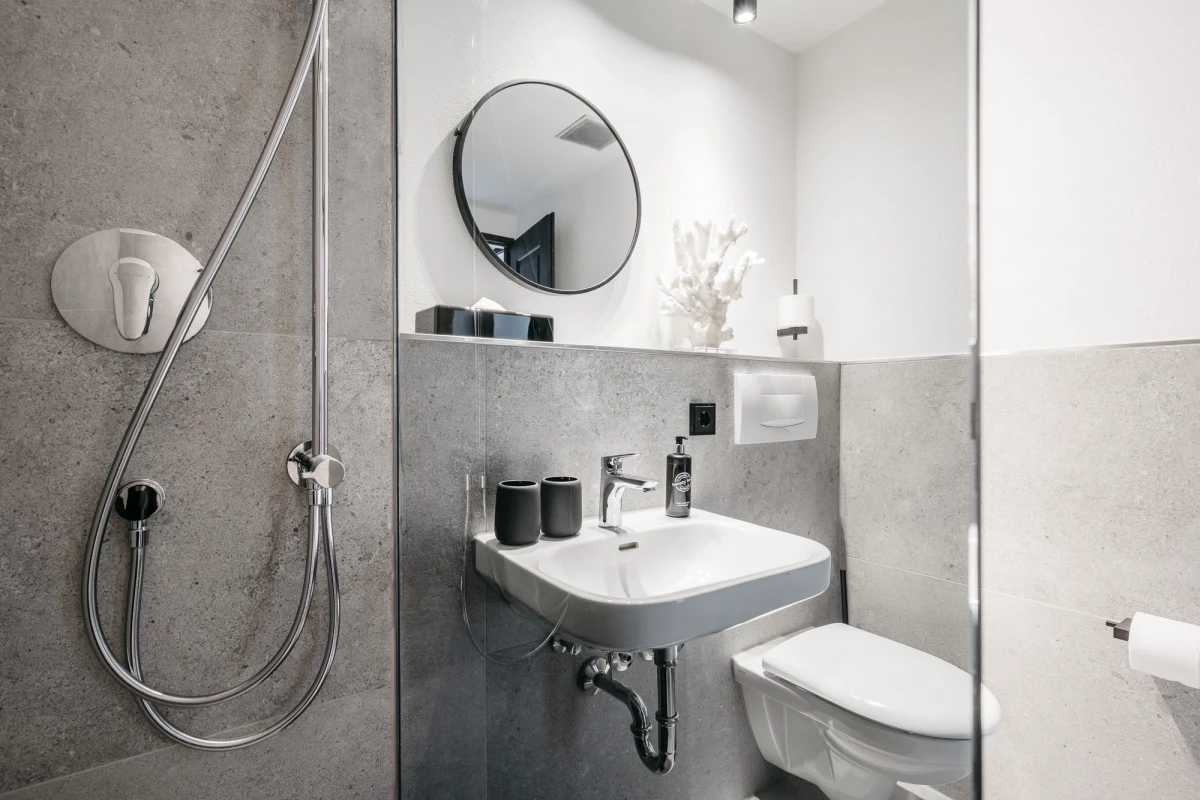 Modernes, minimalistisches Badezimmer mit grauen Betonwänden, weißer Keramik und rundem Spiegel. Ausgestattet mit Dusche, Waschbecken und Toilette in klarer, reduzierter Designsprache.