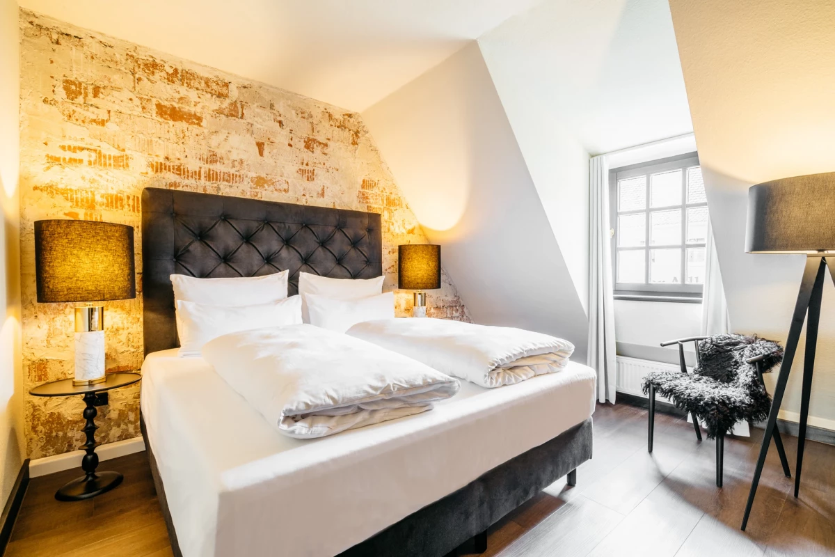Geräumiges Hotelzimmer mit großem gepolstertem Kopfteil und weißer Bettwäsche. Warme Beleuchtung und eine Akzentwand mit vintage Tapete, daneben ein Fenster mit Tageslicht. Ein gemusterter Sessel und eine Stehlampe vervollständigen die einladende Atmosphäre.